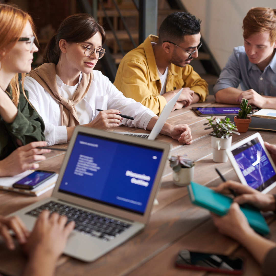 Group of diverse professionals engaged in a collaborative work session around a wooden table, with laptops and digital tablets displaying graphs and notes.
